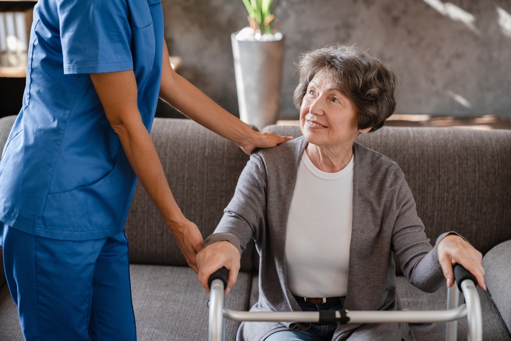 Cropped photo of happy pensioner grandmother elderly patient with walking frame with assistance of nurse caregiver at home. Walking after trauma injury
