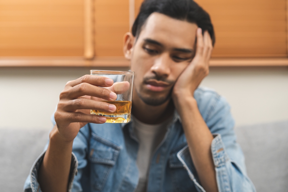 Health care alcoholism drunk, fatigue asian young man hand holding glass of whiskey, alone depressed male drink booze on sofa at home. Treatment of alcohol addiction, suffer abuse problem alcoholism.