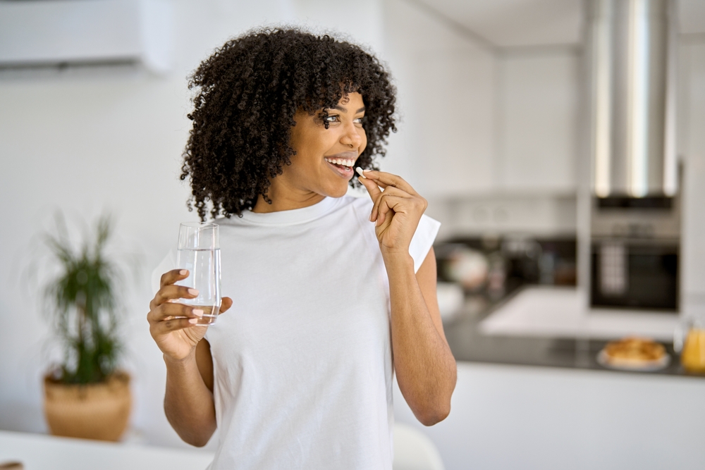 Happy healthy young adult African American woman model talking pill holding glass of water standing in the kitchen at home. Morning supplements vitamins nutrition treatment concept.