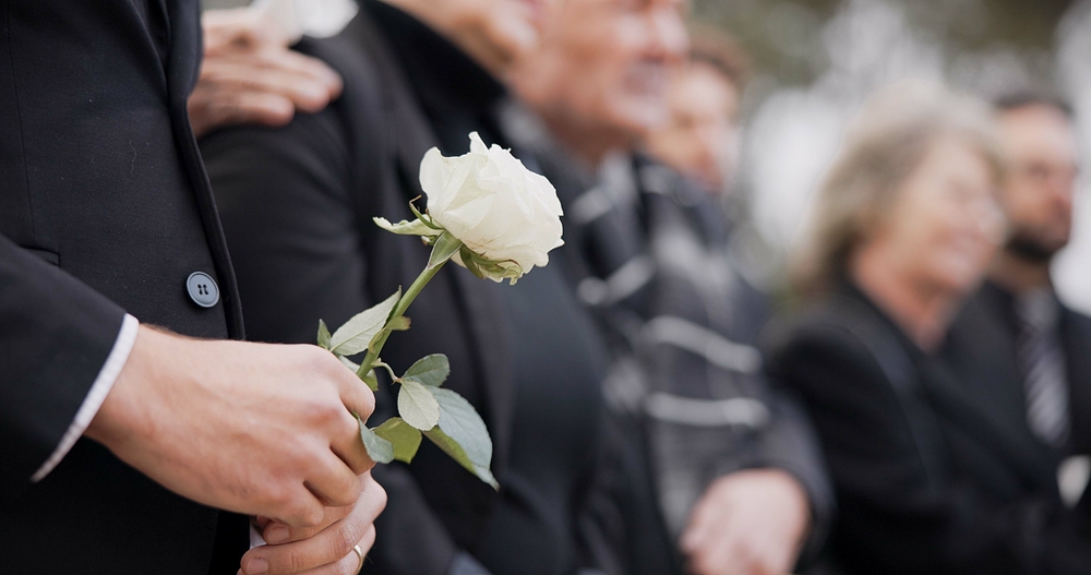 Hands, rose and a person at a funeral in a cemetery in grief while mourning loss at a memorial service. Death, flower and an adult in a suit at a graveyard in a crowd for an outdoor burial closeup
