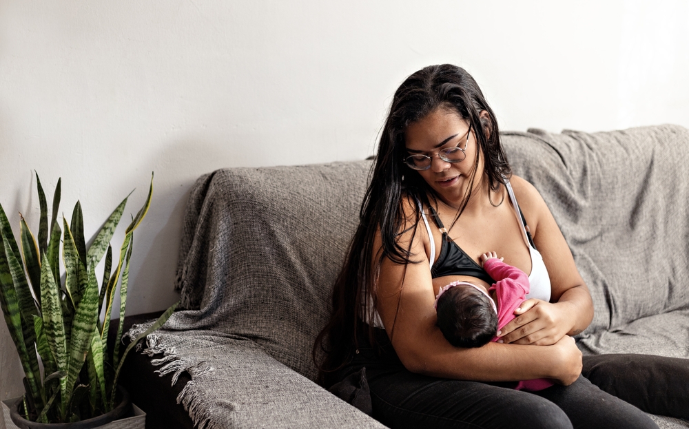 Young mother breastfeeding her beautiful black baby girl. On the couch at her home in Brazil.