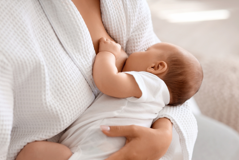 Beautiful young woman in bathrobe breastfeeding her baby at home, closeup