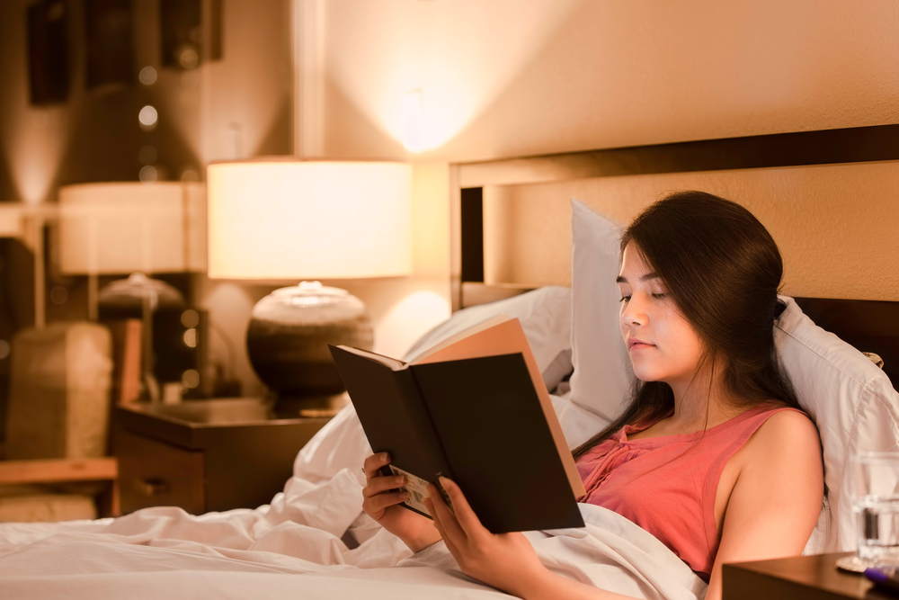 Beautiful biracial Asian Caucasian teen girl reading book in bed at night with yellow lamp light on walls
