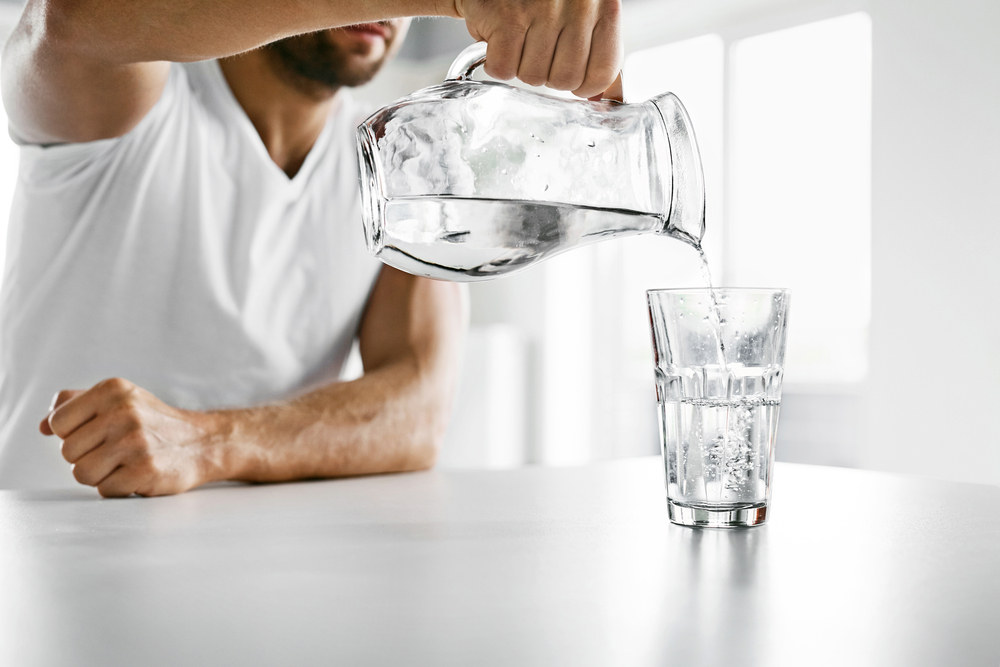 Drink Water. Close Up Of Handsome Young Man Pouring Fresh Pure Water From Pitcher Into A Glass In Morning In Kitchen. Beautiful Athletic Male Model Feeling Thirsty. Healthy Nutrition And Hydration
