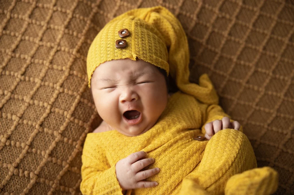 A baby with a yellow onesie and hat, yawning. Brown textured background.