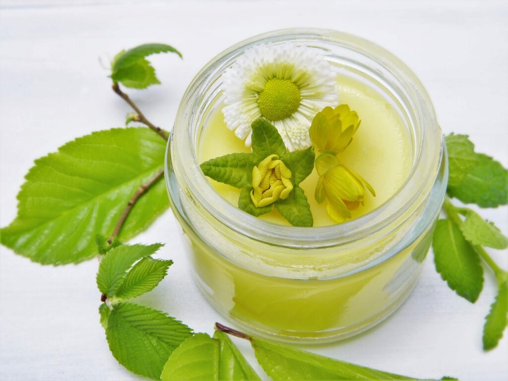 A glass jar of ointment surrounded by blossoms and leaves. White background.