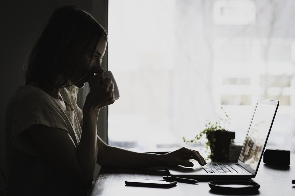 A woman drinking from a mug while sitting at the computer. Desk and window curtains in the background. 