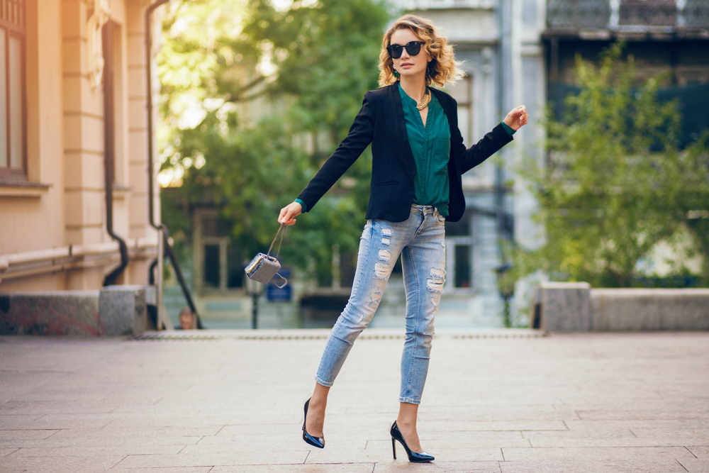 attractive elegant woman walking in city street on high heeled shoes, wearing blue jeans, black jacket, green blouse, sunglasses, holding little purse, fashion trend of summer, slim beautiful lady