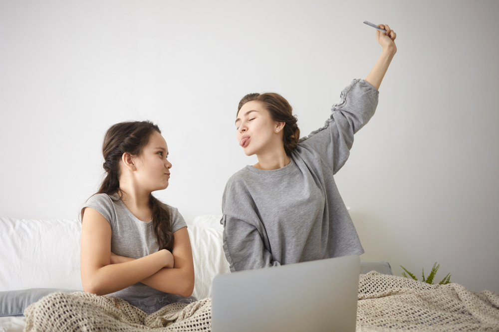 Indoor shot of beautiful casually dressed young female sitting on bed, teasing her angry annoyed daughter, sticking out tongue and taking away credit card while shopping online using laptop