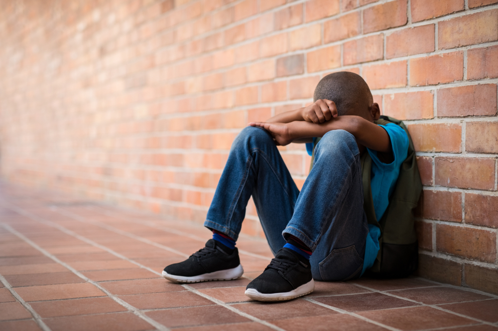Young boy sitting alone with sad feeling at school. Depressed african child abandoned in a corridor and leaning against brick wall. Bullying, discrimination and racism concept with copy space.