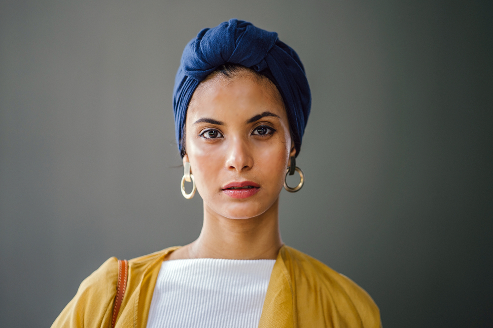 A studio portrait of a young and attractive Muslim Malay woman of Middle Eastern descent. She is stylishly dressed in a yellow cardigan, jeans, white top and a navy turban (hijab head scarf).