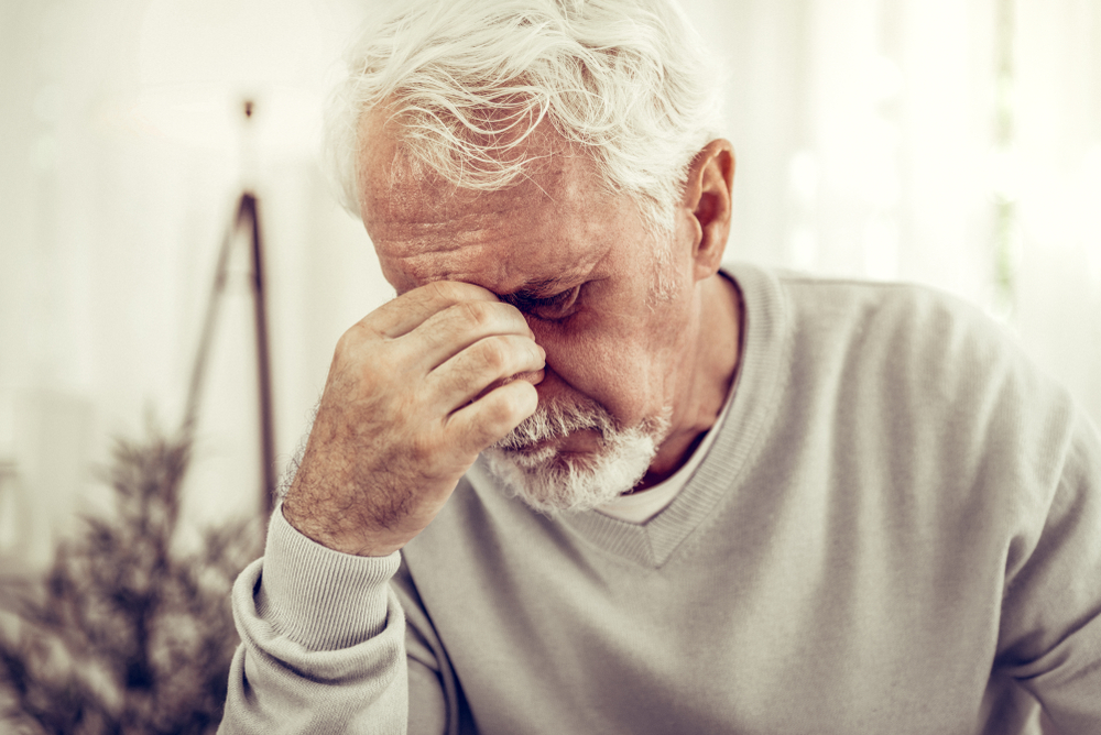 Feeling pain. Old sick grey-haired man in beige sweater feeling terrible pain and keeping his hand on the nose bridge