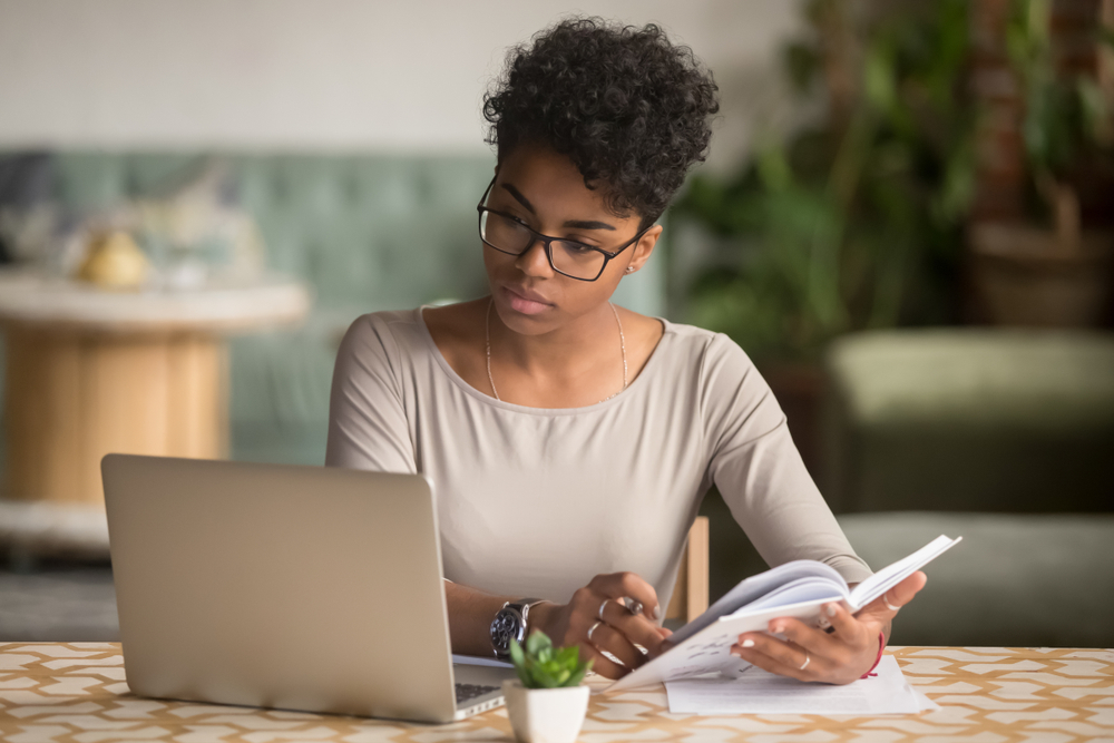 Focused young african american businesswoman or student looking at laptop holding book learning, serious black woman working or studying with computer doing research or preparing for exam online
