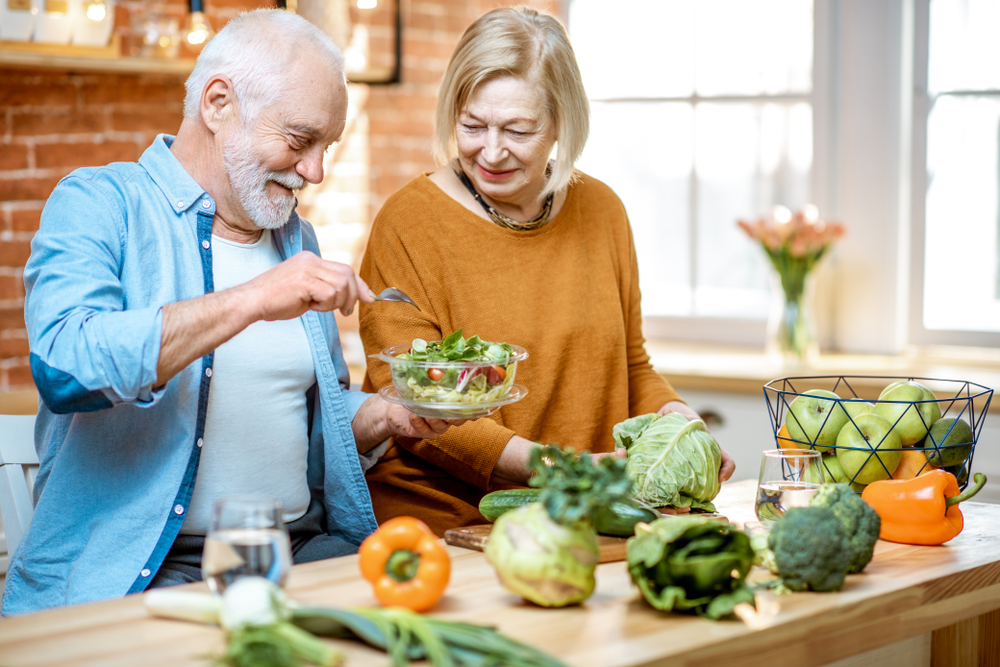 Cheerful senior couple eating salad standing together with healthy food on the kitchen at home. Concept of healthy nutrition in older age
