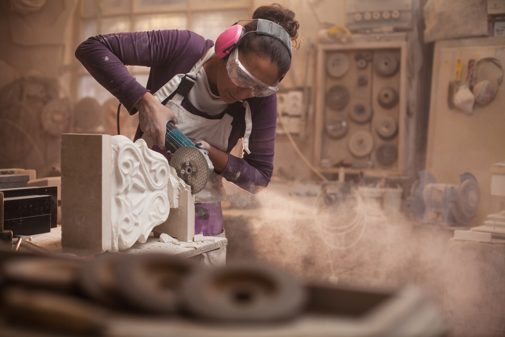 Female stonemason cutting a piece of white marble in a workshop, craftswoman grinding a sculpture with angle grinder, dust and debris flying, women doing hard work concept, stonemasonry and stonecraft