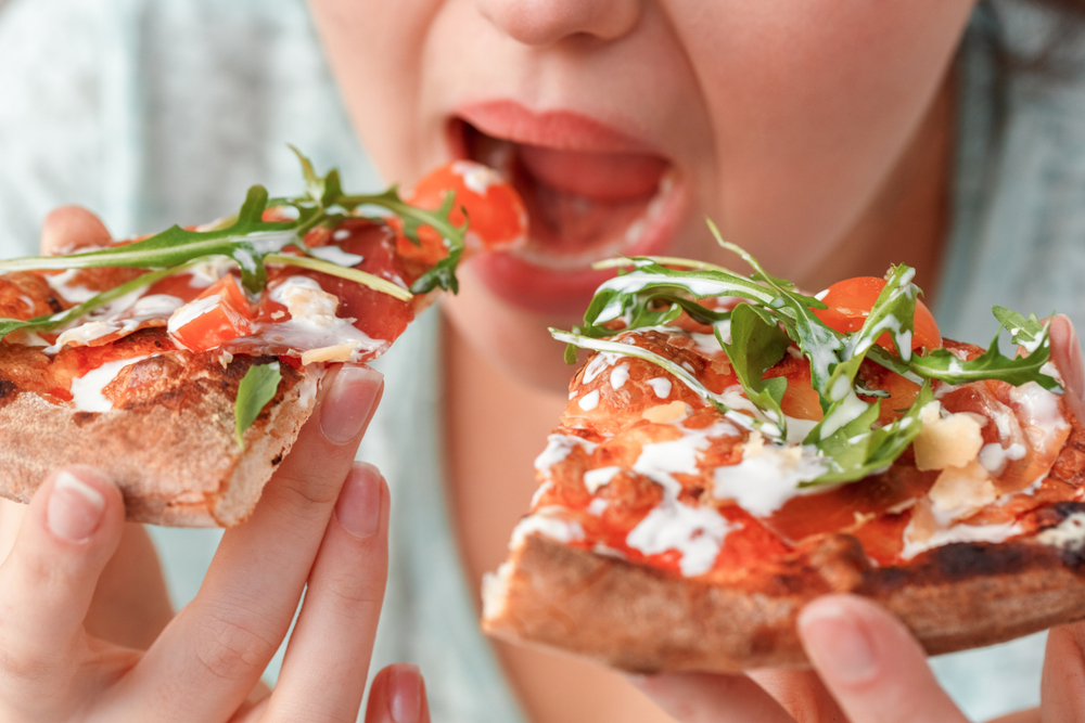 Young chubby woman sitting at table in kitchen binge eating eating slices of pizza fast hungry mouth close-up