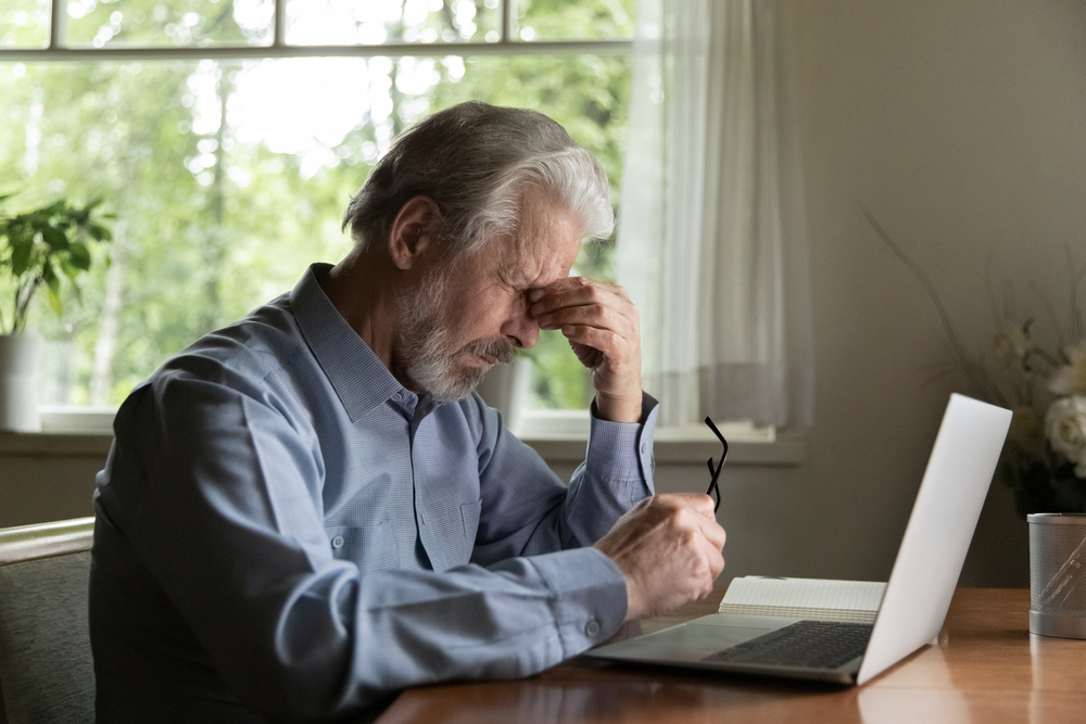 Tired mature 60s man working at laptop from home, taking off glasses, touching head and eyes. Old male pensioner suffering from headache, dizziness after looking at computer screen. Eye sight problem
