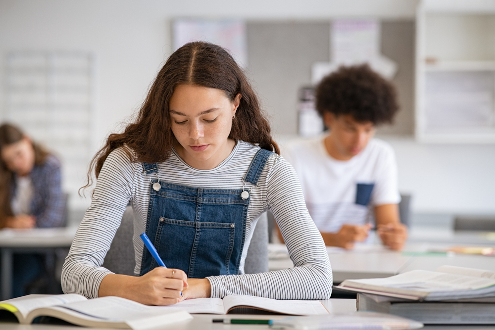 High school student taking notes from book for her study. Young woman sitting at desk and finding information in college library. Focused girl studying in classroom completing assignment.
