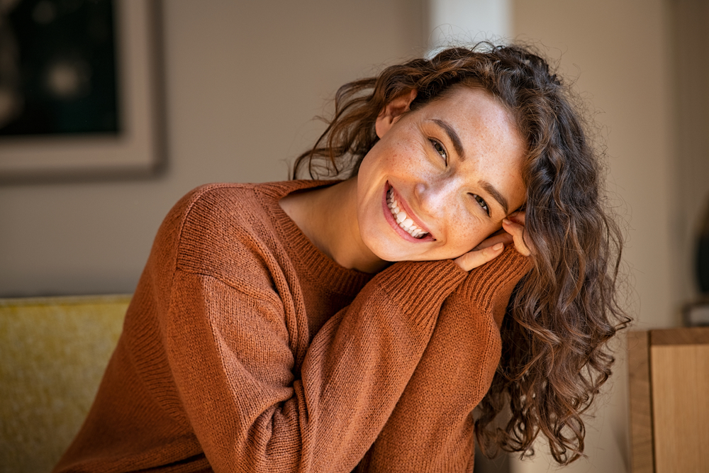 Happy young woman sitting on sofa at home and looking at camera. Portrait of comfortable woman in winter clothes relaxing on armchair. Portrait of beautiful girl smiling and relaxing during autumn.
