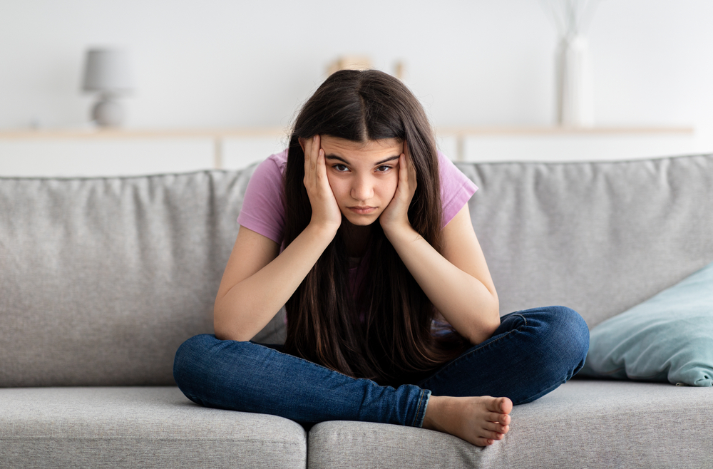 Irritated Indian teenage girl sitting on couch, feeling bored, suffering from lack of communication, being trapped at home during coronavirus lockdown, having teen depression, full length