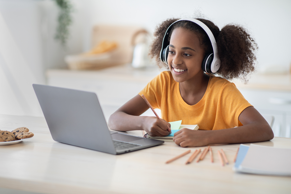 Cheerful black girl studying online, using laptop and headset at home, copy space. Cute african american teen kid having online lesson, drawing, kitchen interior. Home schooling concept