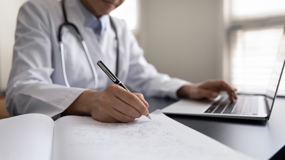 Crop close up Indian woman doctor in white uniform with stethoscope taking notes, using laptop, writing in medical journal, professional therapist practitioner filling documents or patient card
