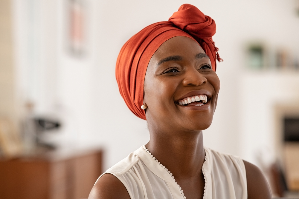 Smiling middle aged african american woman with orange headscarf. Beautiful black woman in casual clothing with traditional turban at home laughing. Portrait of mature carefree lady looking away.
