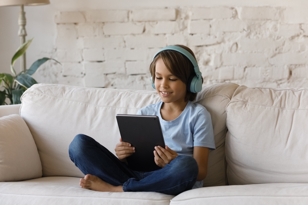 Smiling focused teenage school kid in big wireless headphones using tablet computer, relaxing on sofa at home, watching learning video, movie, attending online class, listening to music