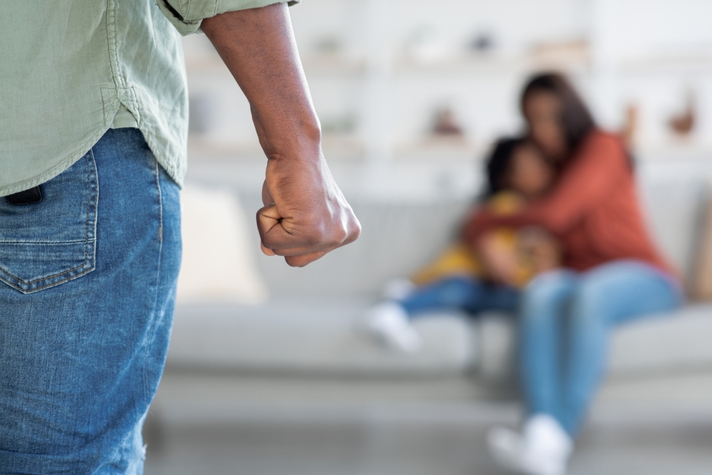 Domestic Violence. Unrecognizable African American Man Threatening Wife And Daughter With His Fist, Scared Mother Embracing Little Girl While Sitting Together On Couch, Selective Focus On Male Hand