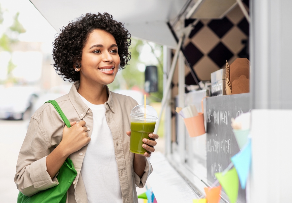 eco living, zero waste and sustainability concept - portrait of happy smiling young woman with green reusable shopping bag drinking takeaway smoothie or shake over food truck background
