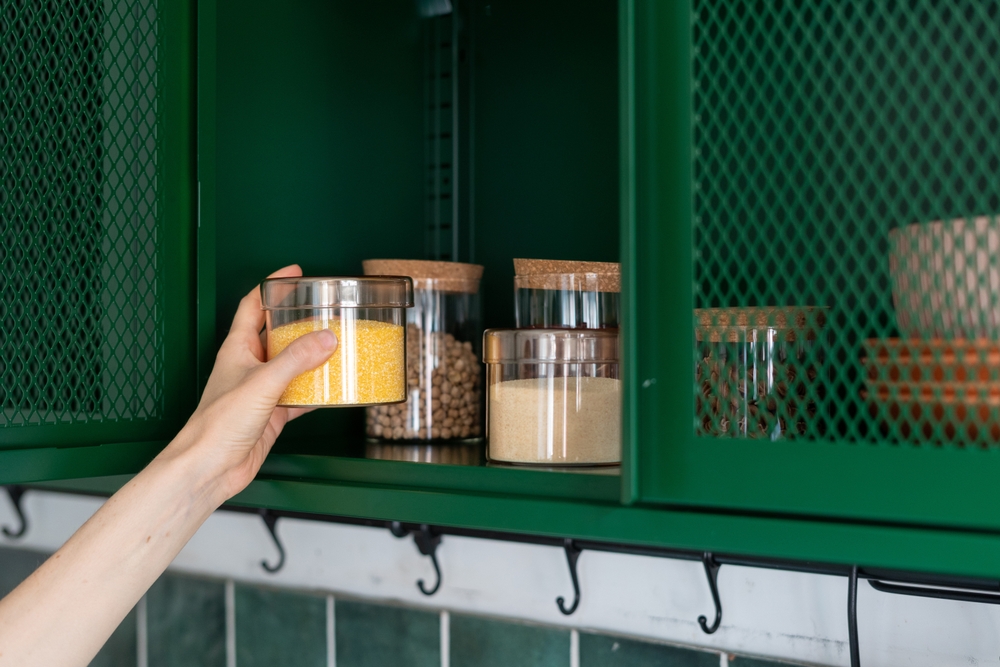 storage grains and beans in jar on shelf in metal green cabinet at kitchen and female hand taking glass container with wheat for prepare food
