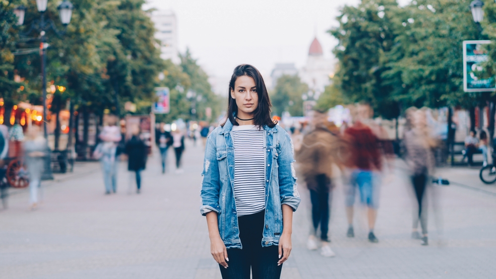 portrait of tired young woman student standing alone in city center and looking at camera with straight face while crowds of men and women are whizzing around.
