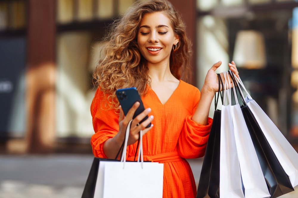 Happy female tourist doing shopping using smartphone while walking in sunny european city. Shopping lifestyle concept, consumerism.