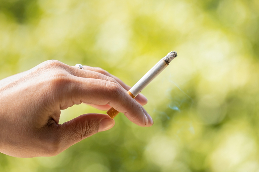 Close up view of smoker holding a cigarette in hand with nature background. Woman with tobacco in her hand surrounded by a natural environment. Cigarette consumed between the fingers. tobacco concept