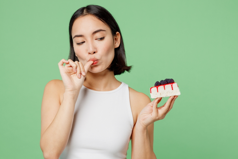 Young happy fun woman wear white clothes holding in hand pice of cake dessert lick fingers isolated on plain pastel light green background. Proper nutrition healthy fast food unhealthy choice concept

