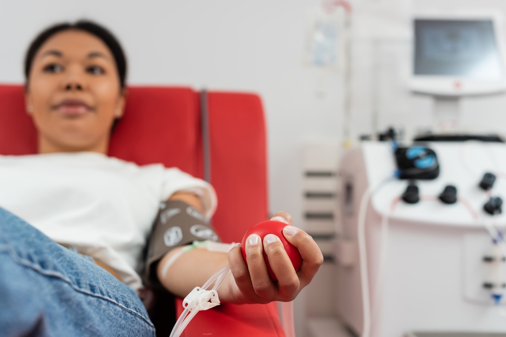 selective focus of medical rubber ball in hand of multiracial woman sitting on comfortable medical chair near transfusion machine while donating blood in laboratory, blurred background