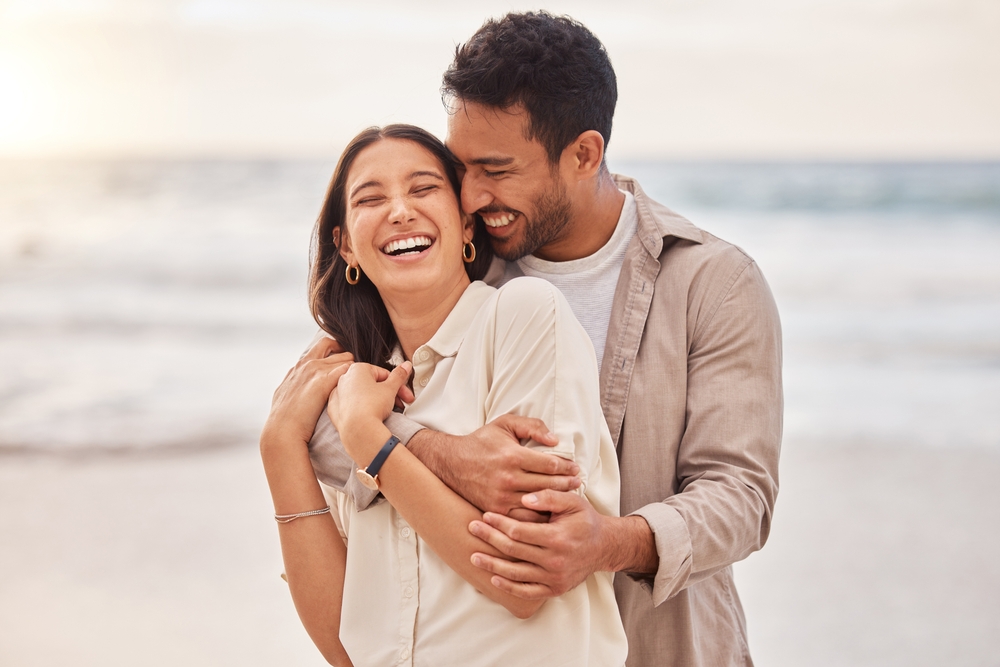 Together, beach and happiness with hug on holiday for having fun with sunset for the weekend. Couple, vacation and laughing with face at the ocean for travel in the summer for a relaxing time.