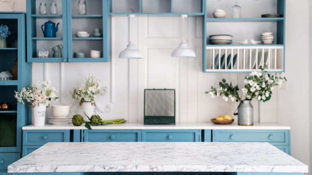 Empty marble kitchen island with clean surface in blue vintage kinchen in provence style. Dining table against blurred stylish furniture with drawers and kitchenware. Pendant lights hanging above.
