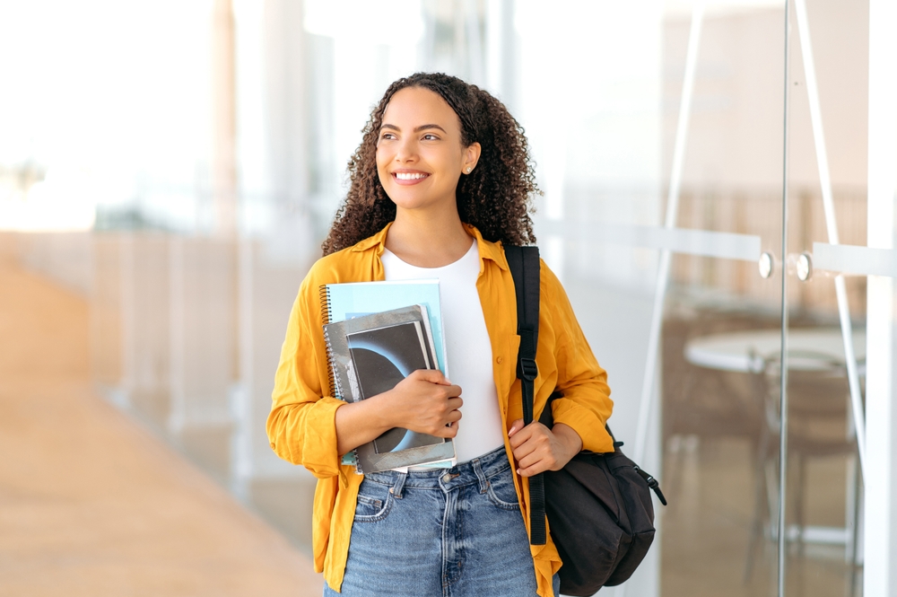 Woman university student. Happy lovely brazilian or hispanic female student, with a backpack, hold books and notebooks in her hand, stand near the university campus, looks and smile to the side
