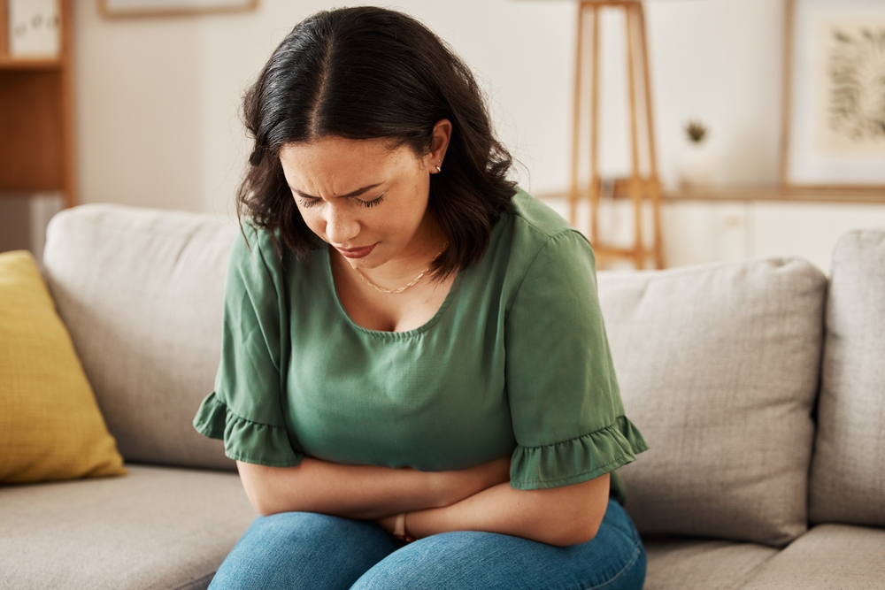 Stress, stomach pain and woman on a sofa with menstruation, gas or constipation, pms or nausea at home. Gut health, anxiety and lady with tummy ache in living room from ibs, bloated or endometriosis
