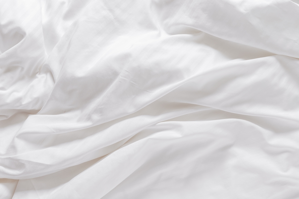 White fabric texture background, wavy cloth. Crease bedclothes fragment as background, Crumpled sheet of Morning bed. White bed linen as minimalistic backdrop. Top view aesthetic surface, horizontal