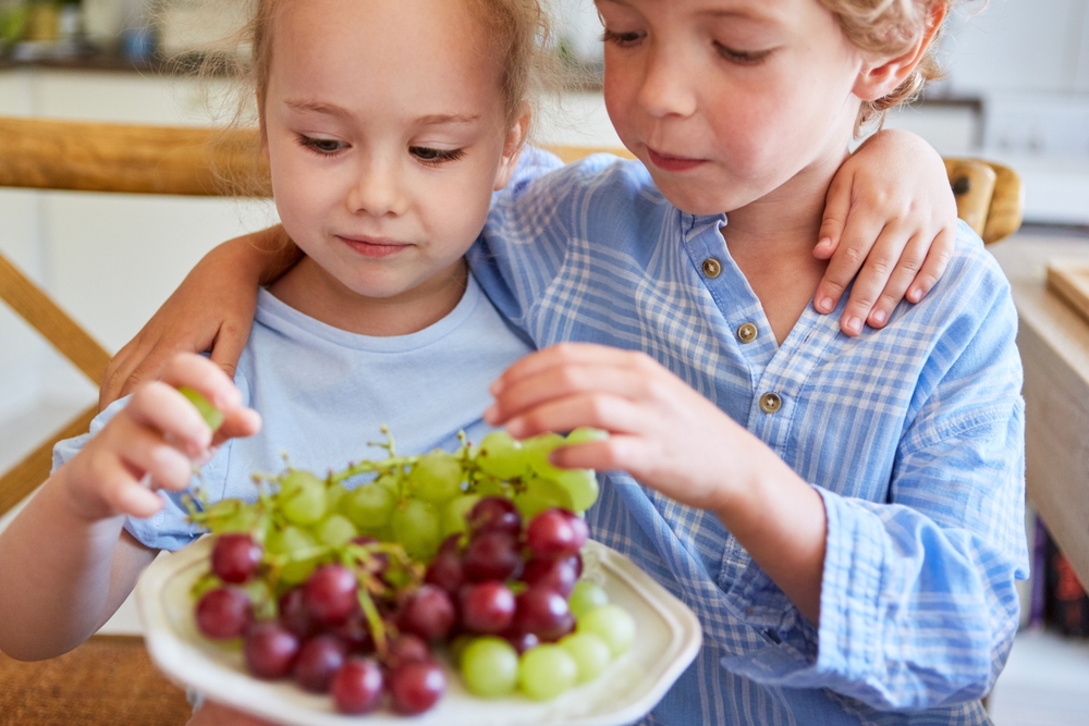 Boy and girl sharing fresh grapes as healthy snack for brother and sister in kitchen
