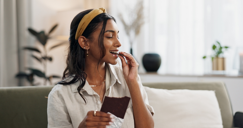 Home, relax and woman with chocolate bar, smile and eating with candy, hungry and dessert. Person, apartment or girl on a sofa, candy or sweet treats with a snack, nutrition or diet plan with cacao
