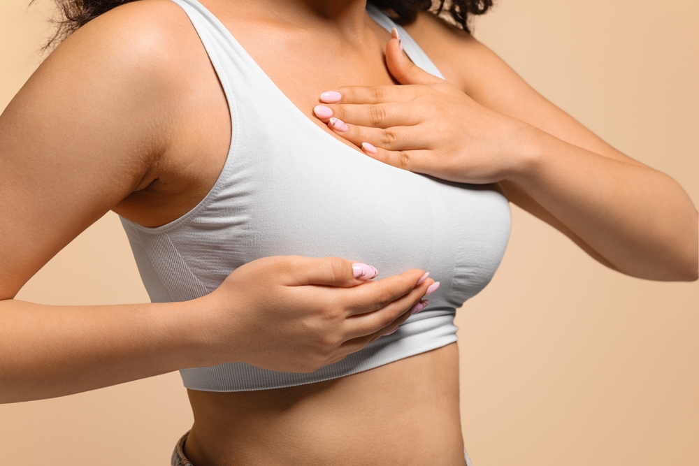 Unrecognizable Young Woman In White Bra Top Hand Checking Lumps On Her Breast, Millennial Female Examining Body For Signs Of Breast Cancer, Standing Isolated On Beige Background, Cropped