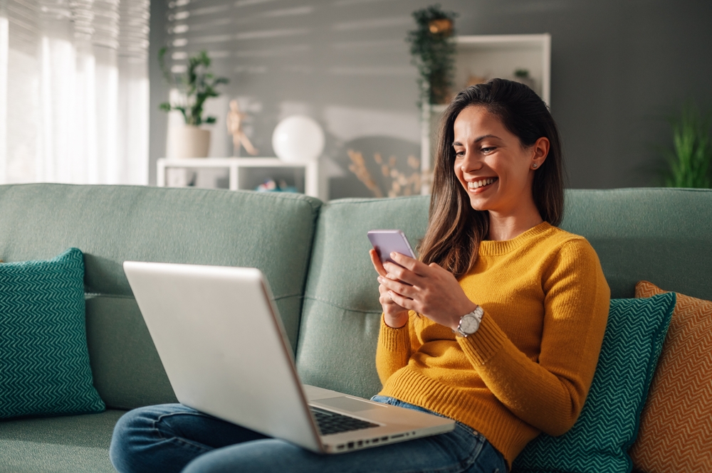 Portrait of a woman using laptop and a phone and checking email or news online while sitting on sofa at home. Searching for friends in internet social networks or working on computer. Copy space.
