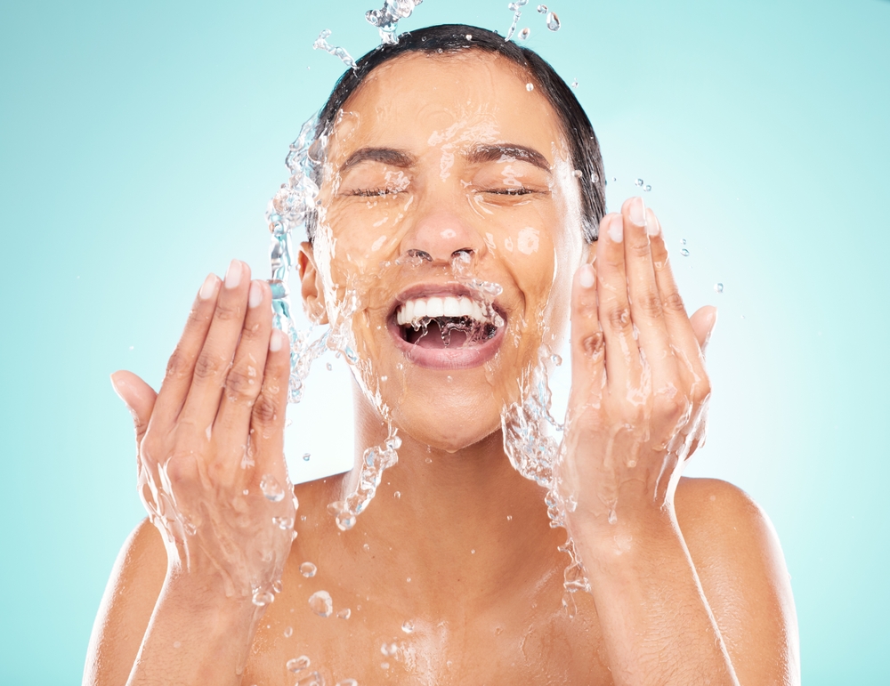 Skincare, water and woman washing face in studio for wellness, hygiene and health isolated on blue background. Beauty, cleaning and excited model splash liquid for dermatology and shower in bathroom
