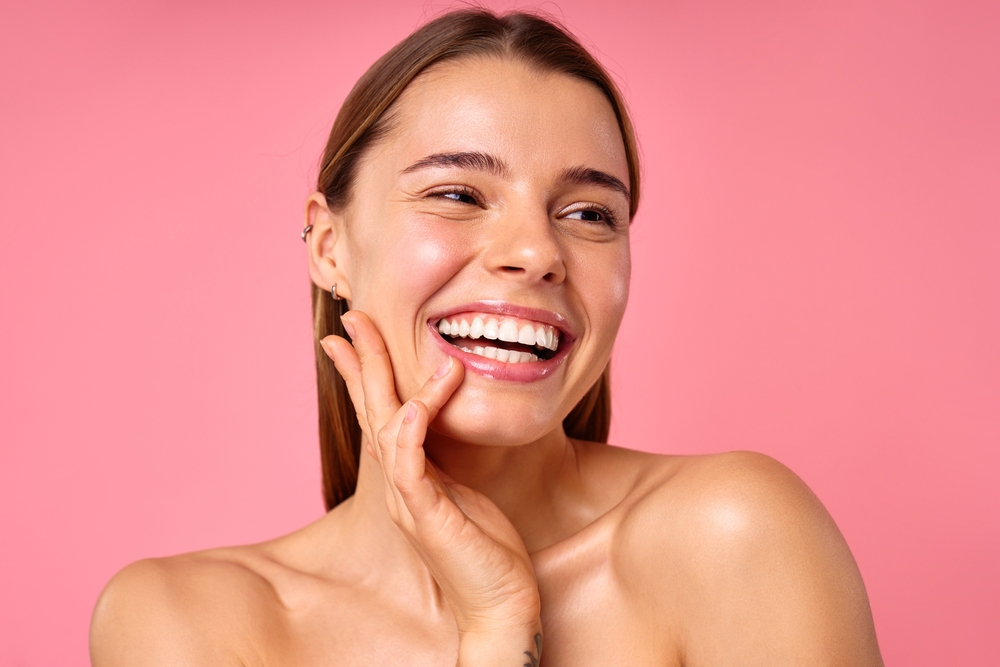 A closeup portrait of a woman with a bright smile, clear and glowing skin, hand on her face on a pink backdrop, showing her natural beauty and confidence, radiating positivity and happiness