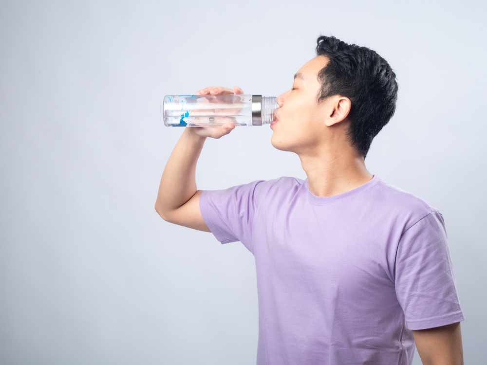 Young Asian man in a light purple shirt, drinking water from a transparent water bottle, standing sideways against a plain light grey background, highlighting hydration and healthy lifestyle.