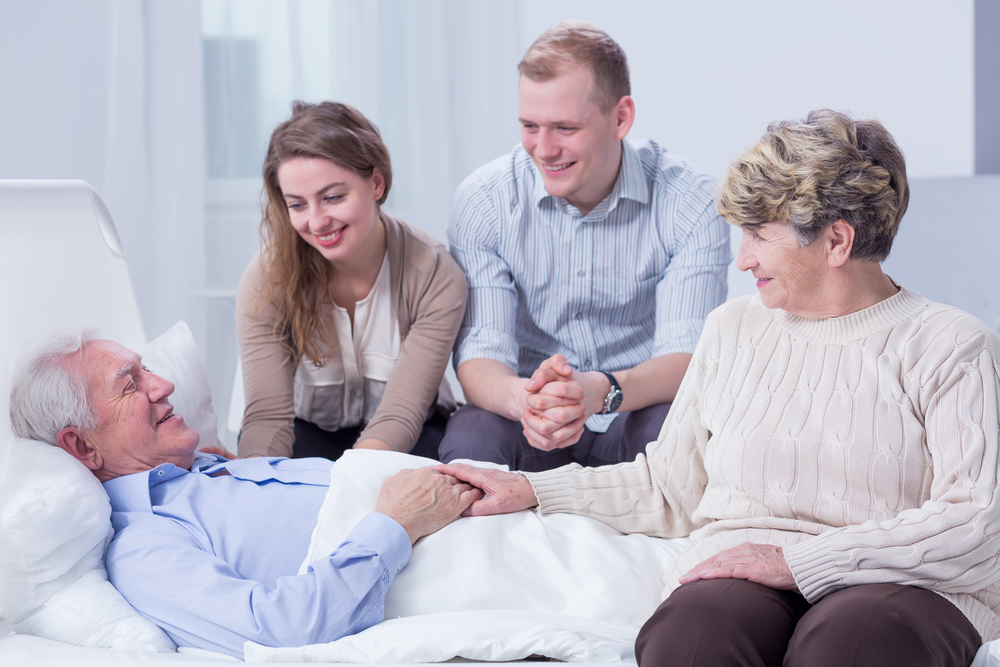 Old man lying in a white hospital bed, surrounded by his smiling relatives
