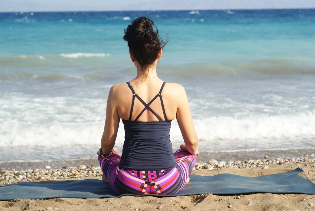 A woman doing yoga on a mat at the beach. Sand and ocean water in the background.